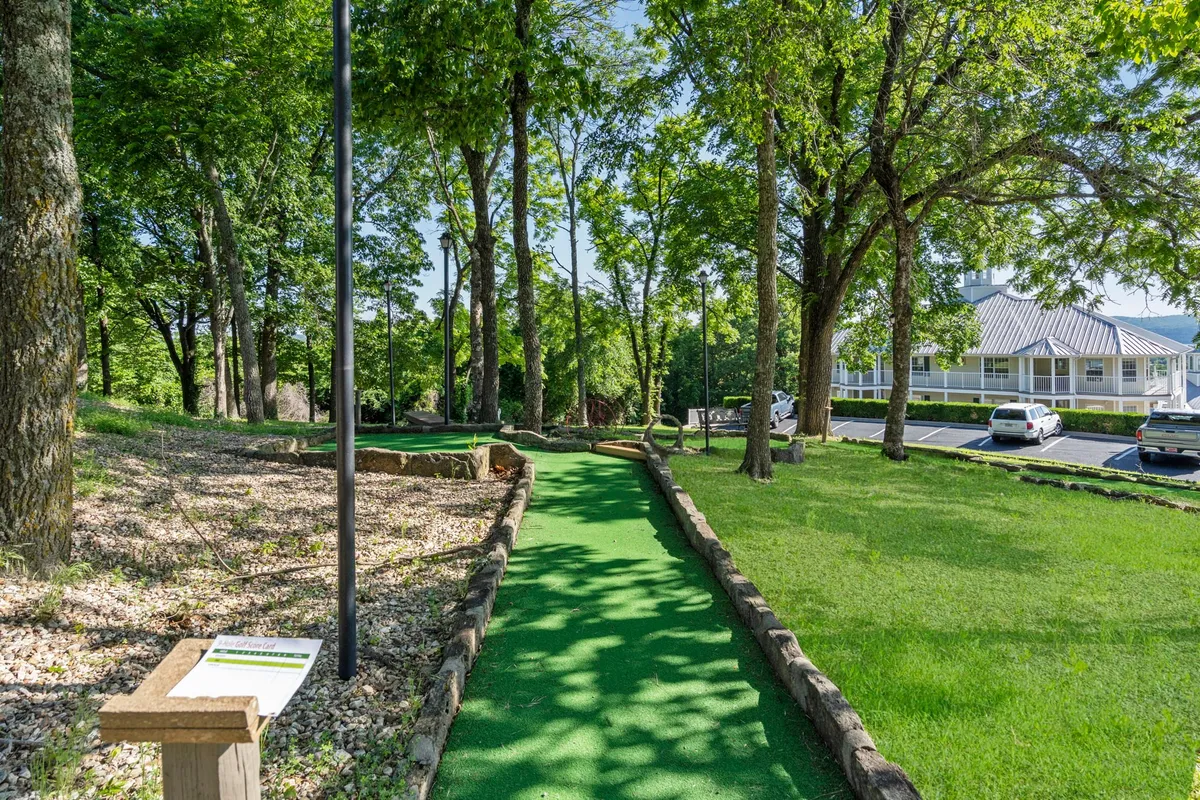 A vibrant exterior view of the Ozark Mountain Resort's mini golf course, surrounded by lush greenery and mature trees. The scene captures children and families enjoying a playful round of mini golf together in a relaxed, natural setting.