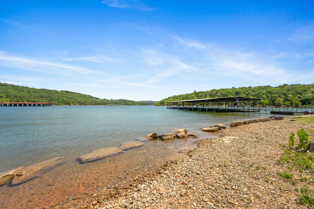 A tranquil lakeside scene featuring calm water, a dock extending into the lake, lush surrounding greenery, and a rocky shoreline. The area offers a peaceful setting ideal for fishing, boating, and enjoying nature.
