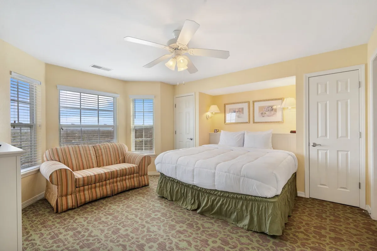 A serene bedroom with a plush bed, a comfortable couch, and large windows allowing ample daylight to flood the space. The setting is calm and inviting, ideal for relaxing, reading, or savoring a peaceful moment.