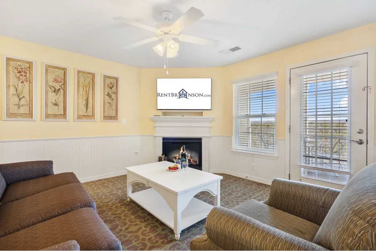A cozy living room scene featuring a plush sofa, coffee table, fireplace, wall art, and TV. The space is awash with natural light, emphasizing the relaxed, inviting atmosphere ideal for gatherings or quiet evenings.