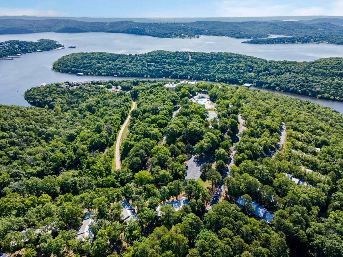This image captures a tranquil lakeside setting with lush greenery, a waterfront view, and scattered homes in the distance. The scene suggests possibilities for walking, hiking, and relaxing by the water's edge with ample natural shade.