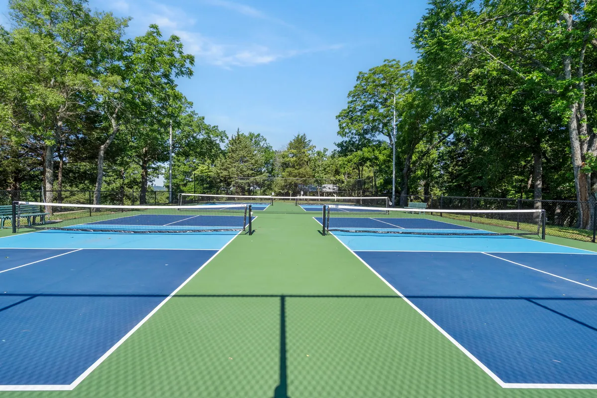 Energetic tennis courts surrounded by green foliage, with clear netting and open sky above. The setup suggests both friendly competition and a connection to the outdoors.